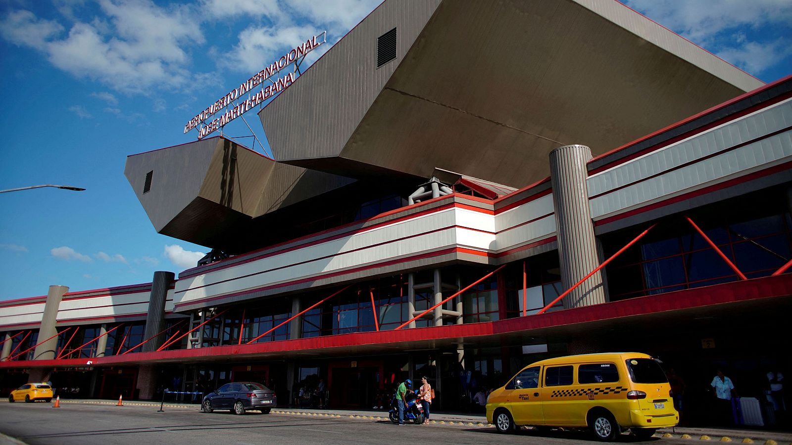 En el Aeropuerto Internacional, se aprecia un edificio de diseño moderno con una estructura geométrica singular. Hay vehículos y personas, indicando actividad en el lugar.