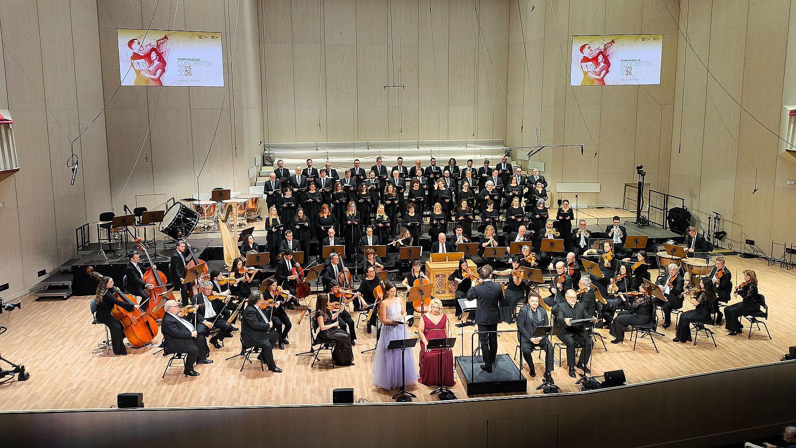 Una orquesta y un coro, ambos vestidos de negro, actúan en un escenario iluminado. El director dirige a los músicos de la orquesta, que tocan instrumentos variados. Detrás, el coro participa en la interpretación, con pantallas proyectando imágenes.