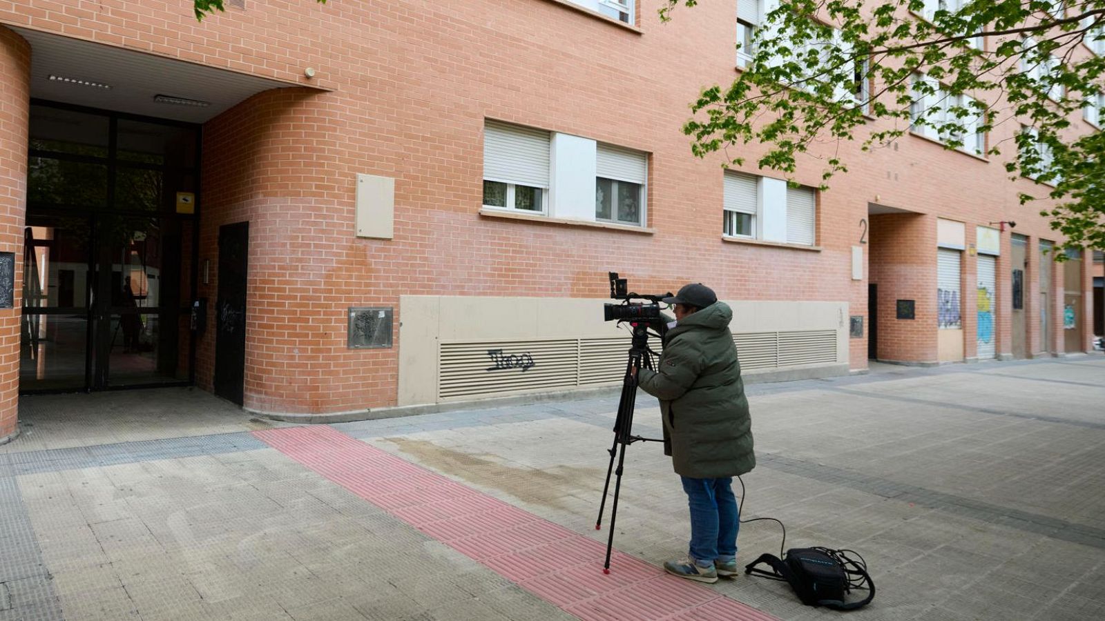 Un cámara, con chaqueta oscura y gorro, graba con una cámara profesional en un entorno urbano con un edificio de ladrillo rojo como telón de fondo, posiblemente en un barrio de Pamplona.