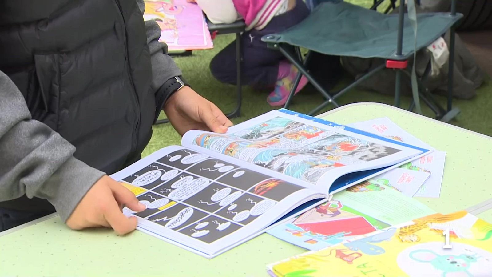 En un evento al aire libre, una persona lee un cómic abierto, mostrando páginas con viñetas y bocadillos de diálogo. La mesa, rodeada de libros y papeles, sugiere un ambiente relajado.