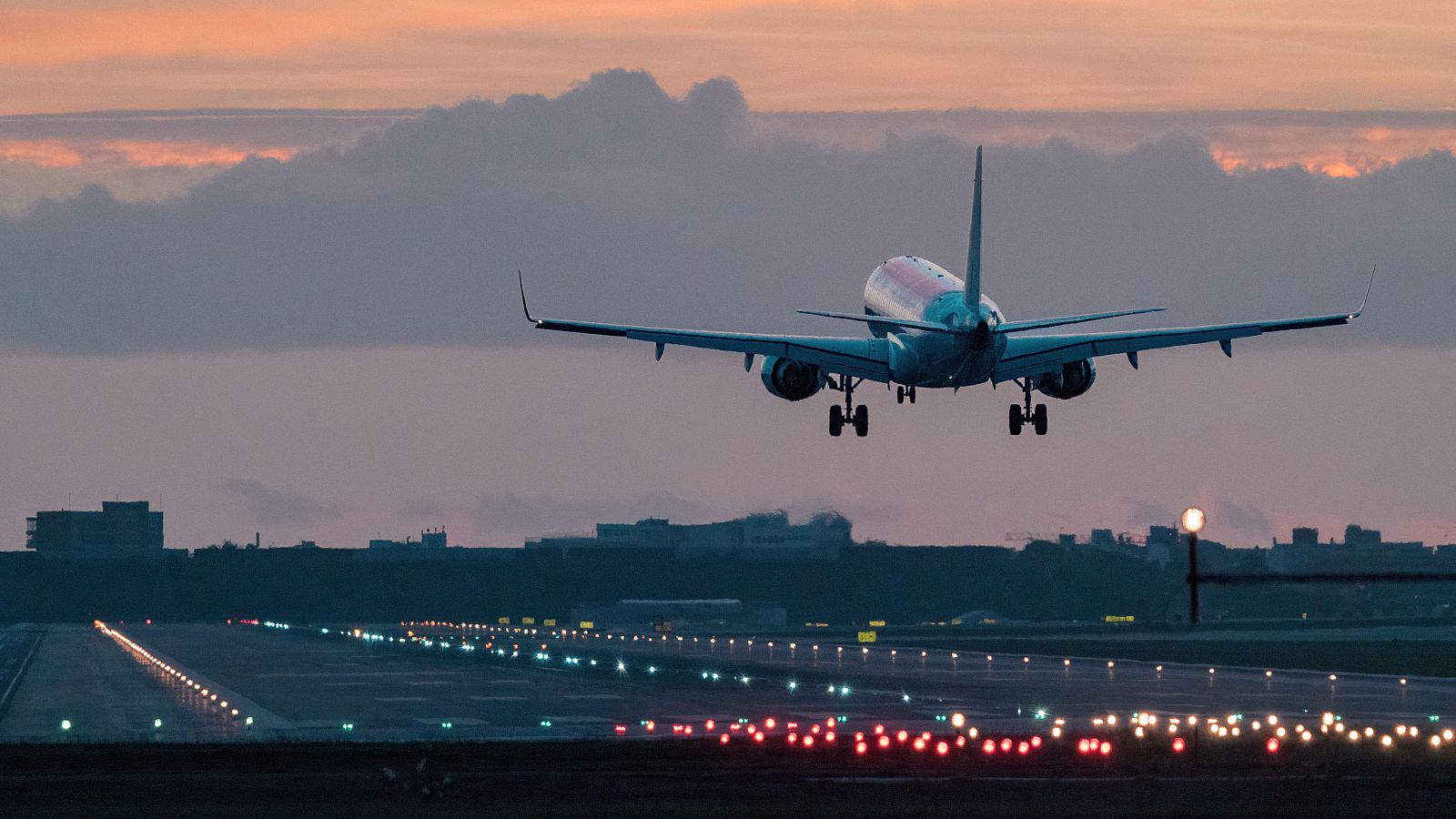 Un avión de pasajeros, con detalles en rojo y blanco, se aproxima a la pista de aterrizaje iluminada, con un cielo teñido de tonos rosados y anaranjados, posiblemente al amanecer o atardecer, y un horizonte urbano al fondo.