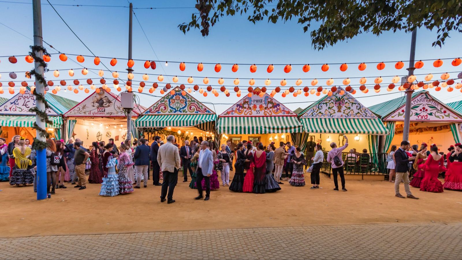 Una vista general de la Feria de Abril revela casetas adornadas con toldos a rayas y nombres, como "El Glorioso Rege", y personas vestidas con trajes tradicionales, disfrutando del ambiente festivo.
