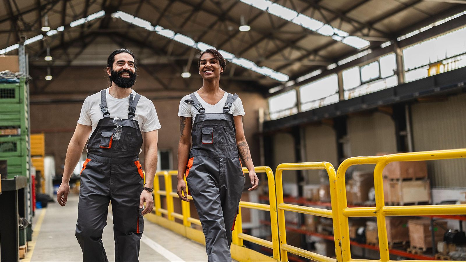 Un hombre y una mujer de origen extranjero, vestidos con ropa de trabajo, sonríen en el interior de una nave industrial