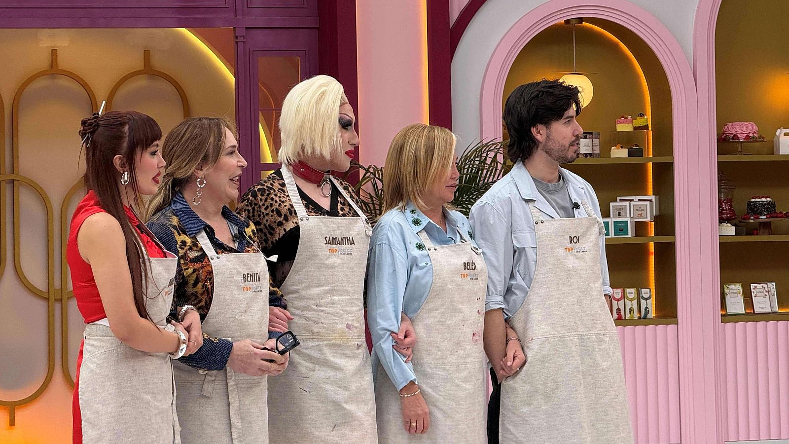 Cinco personas, incluyendo a una drag queen, posan en un set de televisión. Todos visten delantales con nombres y el fondo presenta una estantería con dulces y una decoración en tonos rosa y dorado.