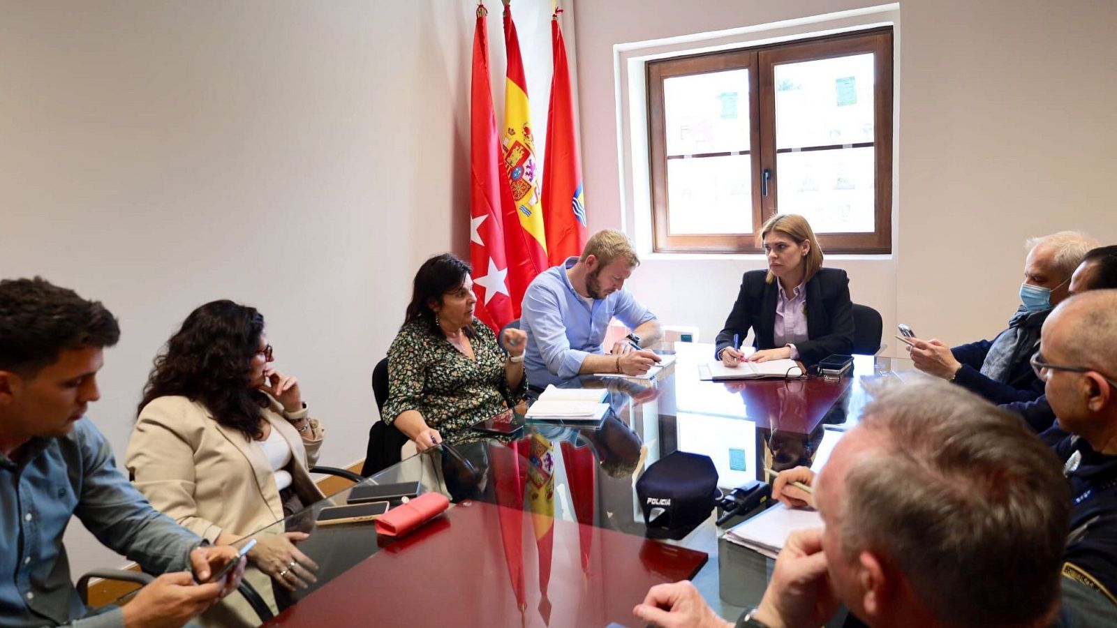 En una sala de reuniones, varias personas están sentadas alrededor de una mesa roja, con documentos y teléfonos. Se distinguen banderas y asistentes tomando notas y usando mascarillas.
