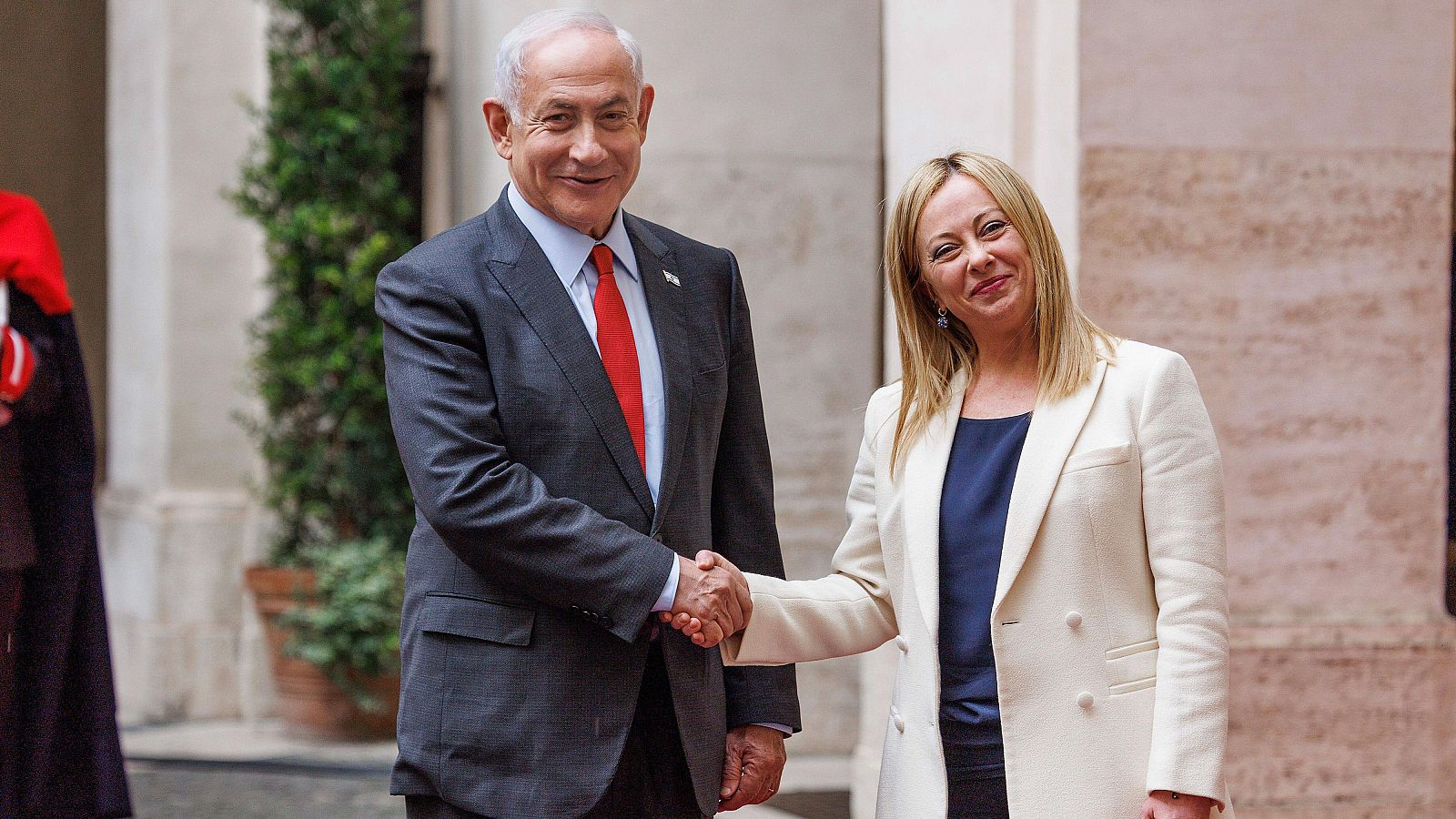 En una reunión oficial, Giorgia Meloni y Benjamin Netanyahu se saludan. Netanyahu, con traje oscuro y corbata roja, y Meloni, con chaqueta clara, se encuentran en un espacio que sugiere un edificio gubernamental.