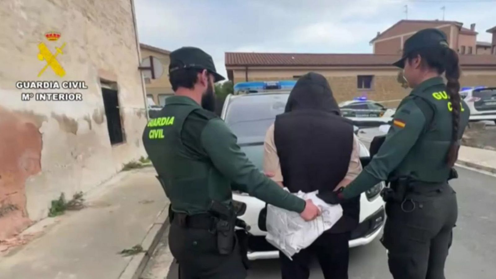 Dos agentes de la Guardia Civil uniformados custodian a una persona arrestada que lleva una sudadera con capucha, un chaleco negro, esposas y una bolsa blanca.