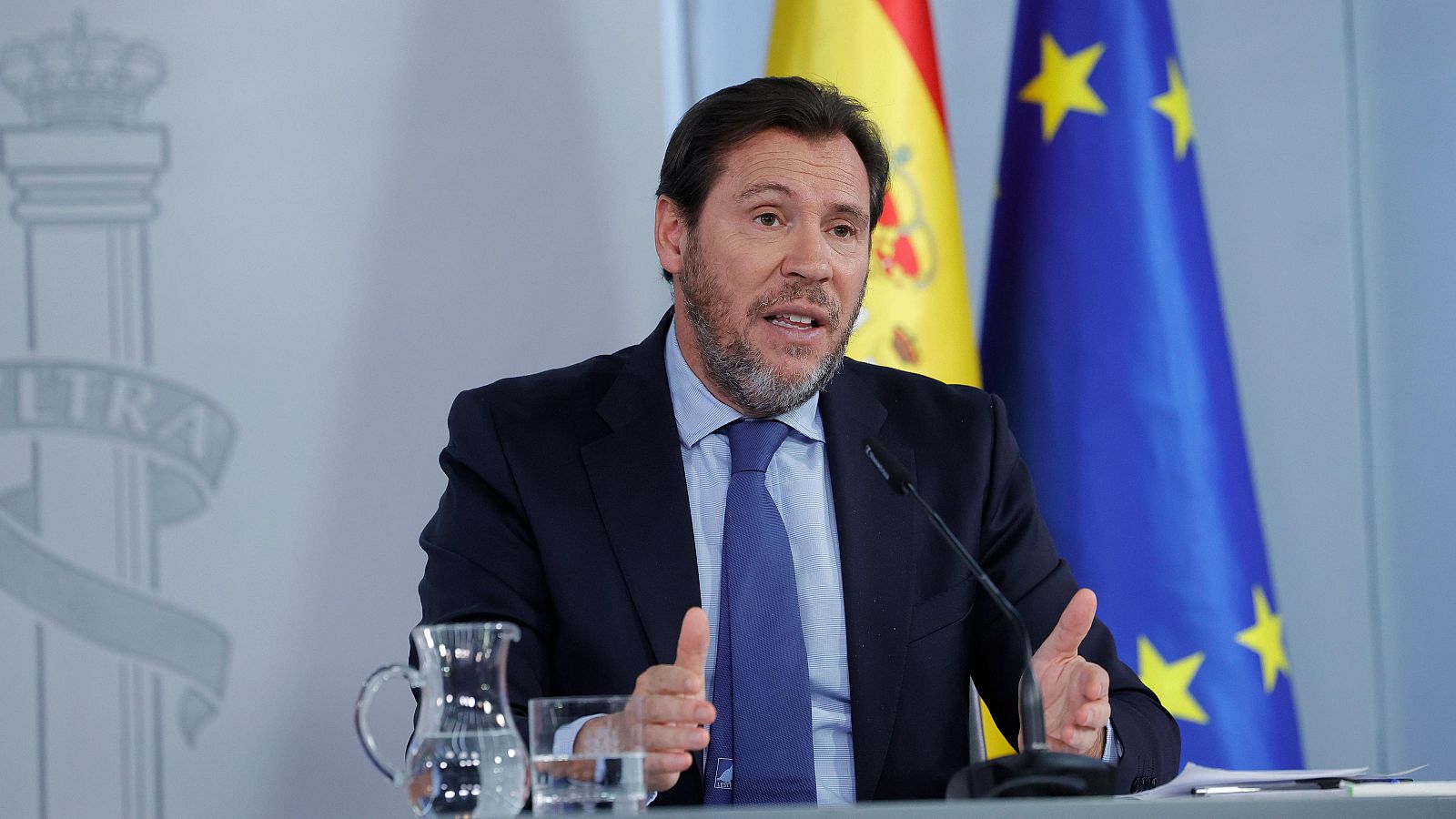 Un hombre de mediana edad, con barba y traje oscuro, se encuentra frente a un micrófono, con banderas de España y la Unión Europea de fondo. Hay una jarra de agua sobre la mesa.