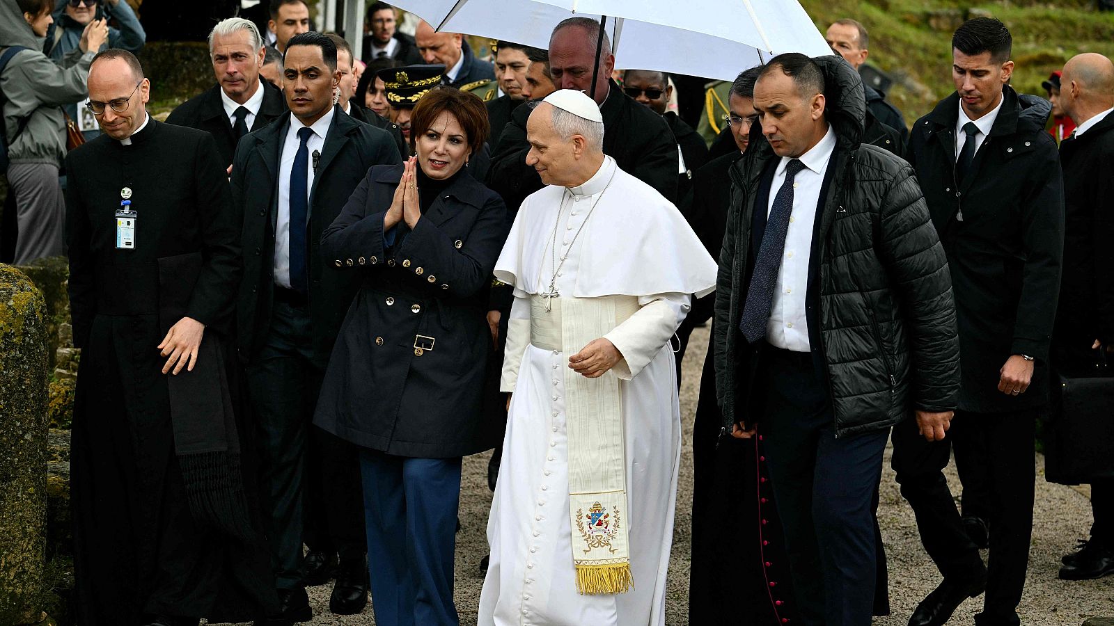 El Papa Francisco, vestido con su atuendo blanco, camina en un sitio arqueológico junto a una mujer. Están rodeados de personas, incluyendo personal de seguridad y sacerdotes, con restos de estructuras antiguas visibles.