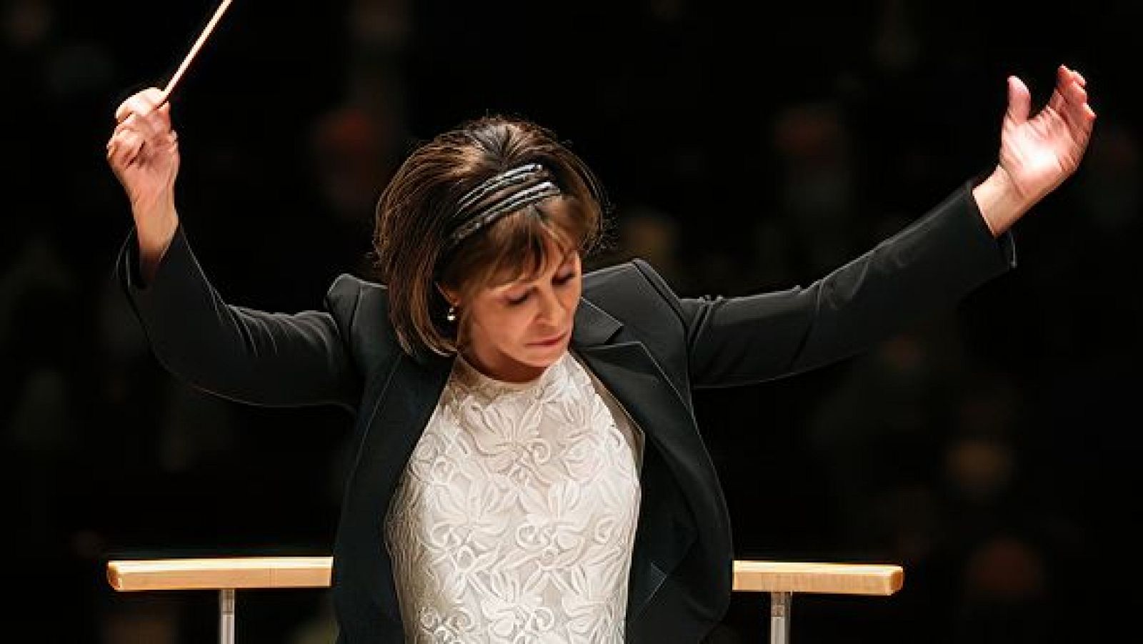 Una directora de orquesta, con diadema plateada y vestimenta elegante, dirige a un conjunto musical, probablemente una orquesta y un coro, con una batuta en la mano, en un espacio con fondo oscuro.