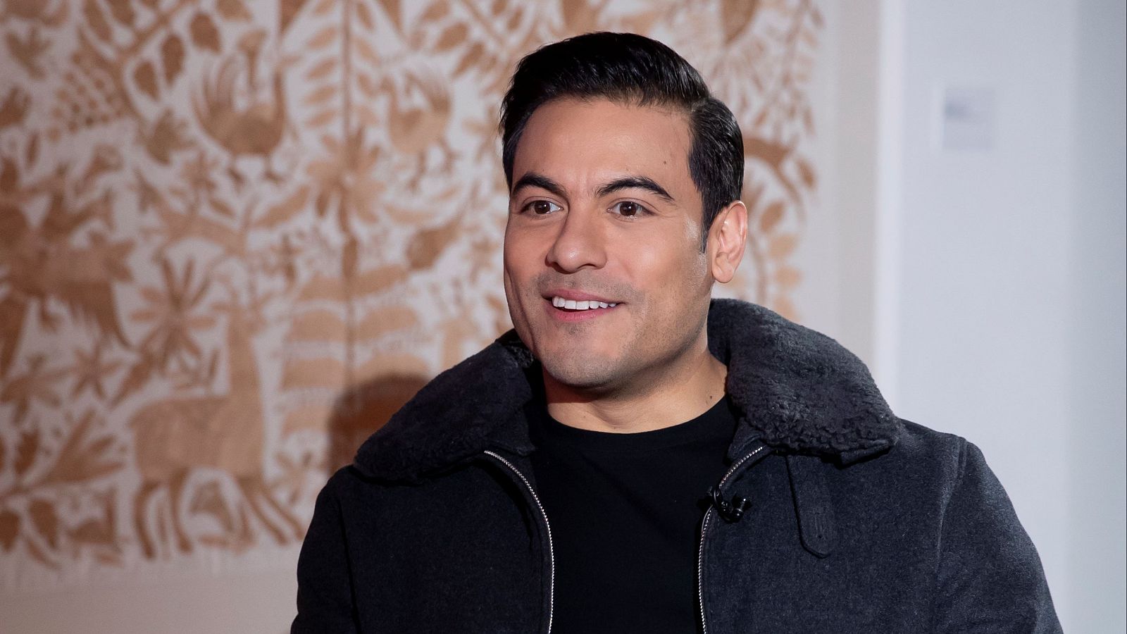 En un plano general, Carlos Rivera sonríe, luciendo una camiseta negra y una chaqueta oscura con cuello de borrego, con el cabello peinado hacia atrás. El fondo presenta una pared con un diseño de animales y flores en tonos claros.