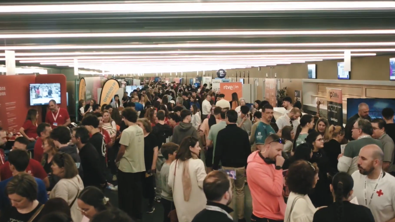 En un evento concurrido, se observa una multitud interactuando en un espacio amplio con stands y pantallas. Algunas personas llevan identificaciones, y se distinguen logotipos como el de una cadena de televisión.