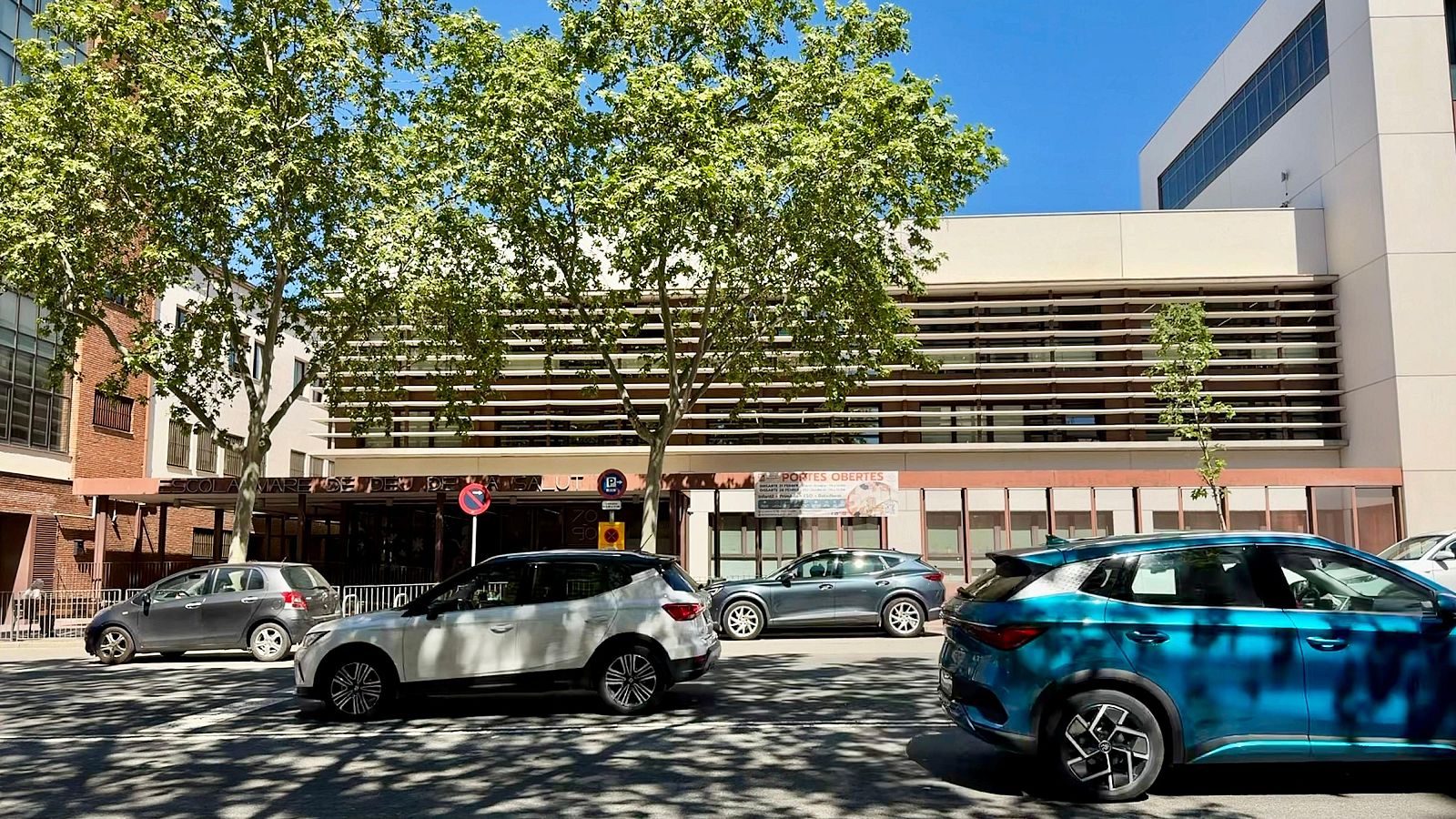 Una vista de la Gran Via de Sabadell amb cotxes en moviment davant d'un edifici modern, possiblement un centre educatiu, amb arbres i senyals de trànsit.
