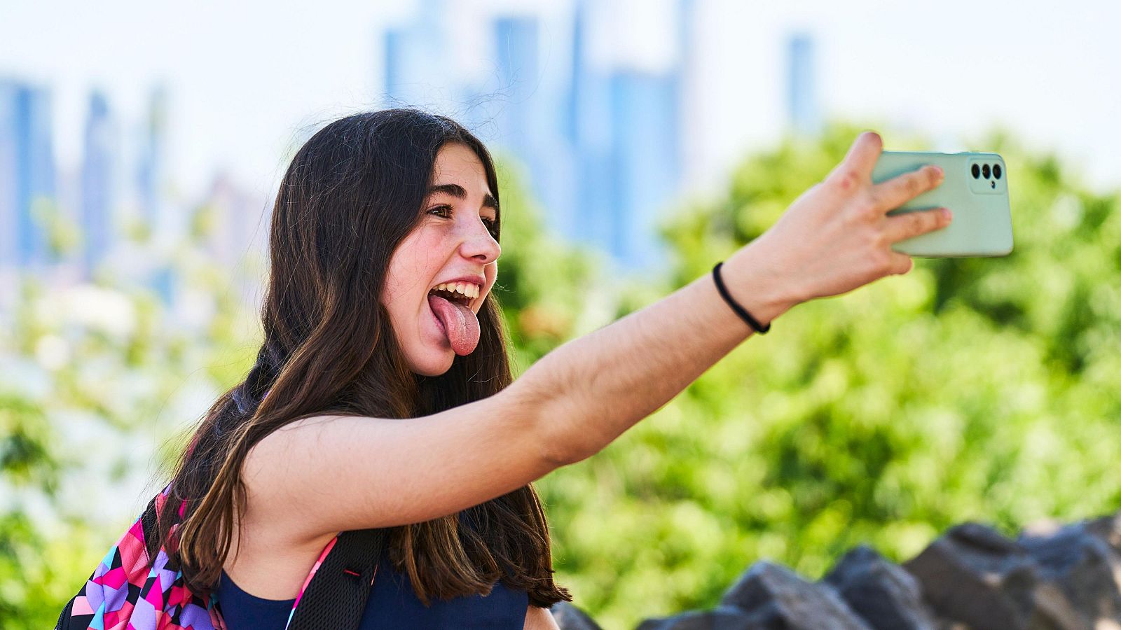 Una adolescente sonríe a su teléfono móvil en un parque soleado, transmitiendo una sensación de bienestar y conexión social.