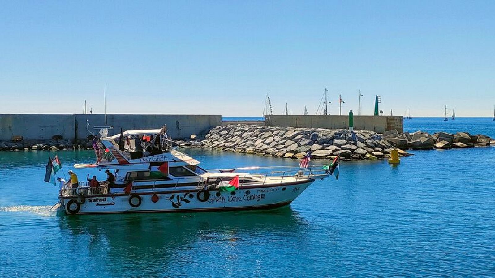 Un barco blanco, identificado como "Safad", navega con banderas palestinas y estadounidenses, llevando el mensaje "Free Gaza" y "Gaza we're coming!!". Se observa a personas a bordo, con un puerto y otros barcos al fondo, bajo un cielo azul.