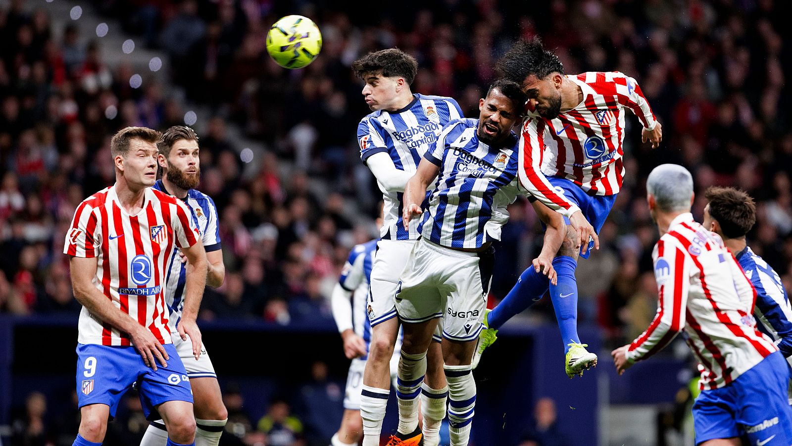 Un partido de fútbol enfrenta al Atlético de Madrid y a la Real Sociedad, con jugadores disputando el balón en el aire, en un estadio con público.