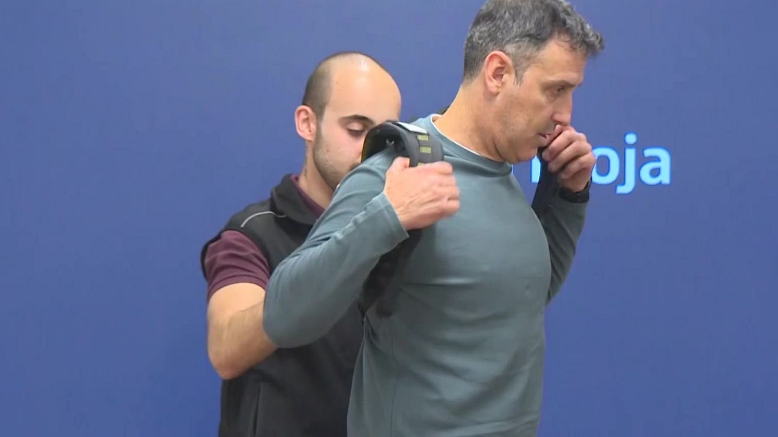 Un hombre con camiseta burdeos y chaleco negro asiste a otro, de pelo canoso y camiseta verde azulado, a colocarse un dispositivo. El fondo es de color azul.