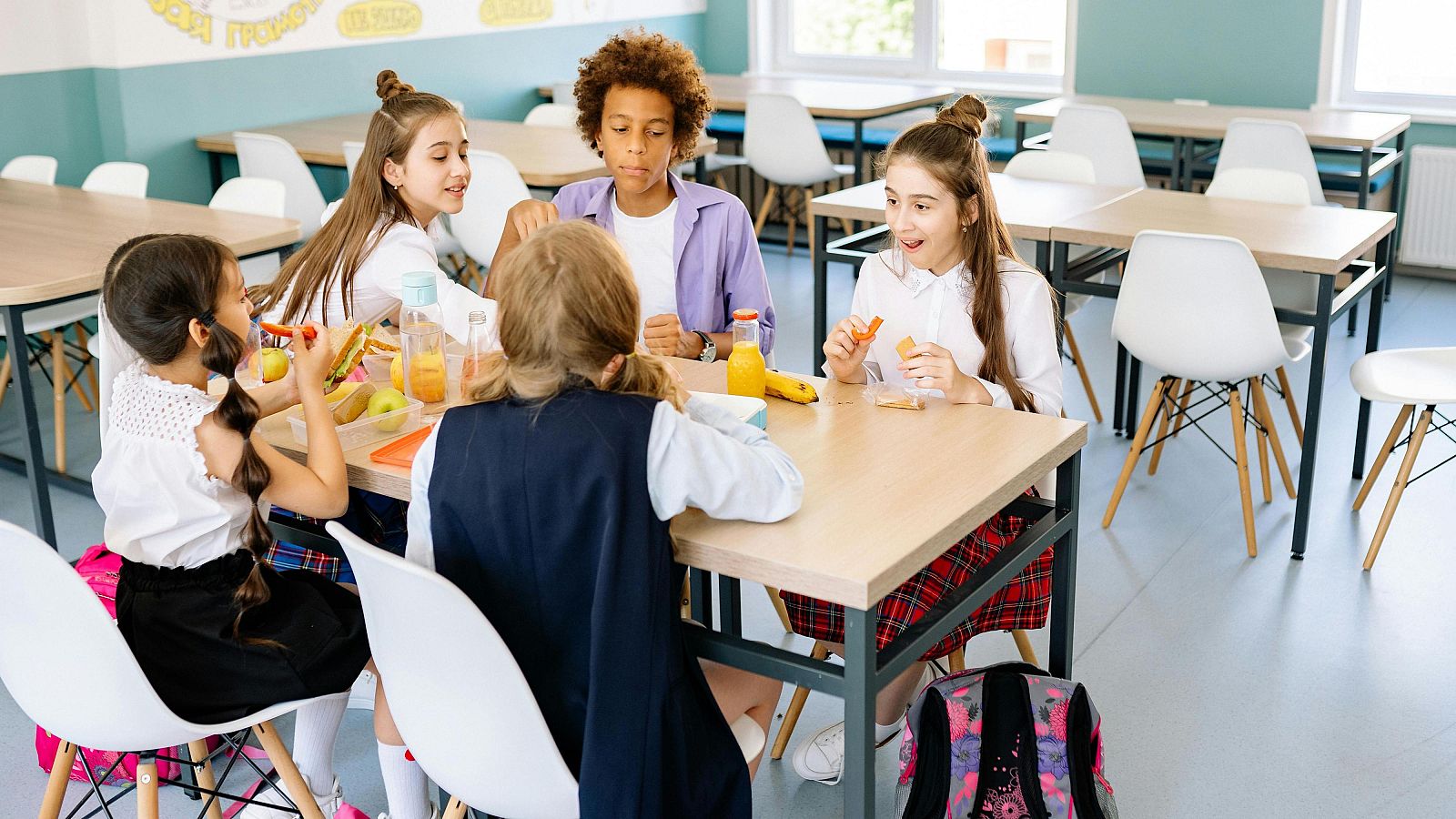 En un comedor, un grupo de niños de diferentes orígenes étnicos están sentados alrededor de una mesa comiendo. Se observan alimentos como zumo, frutas y sándwiches, y algunos niños visten uniformes escolares.