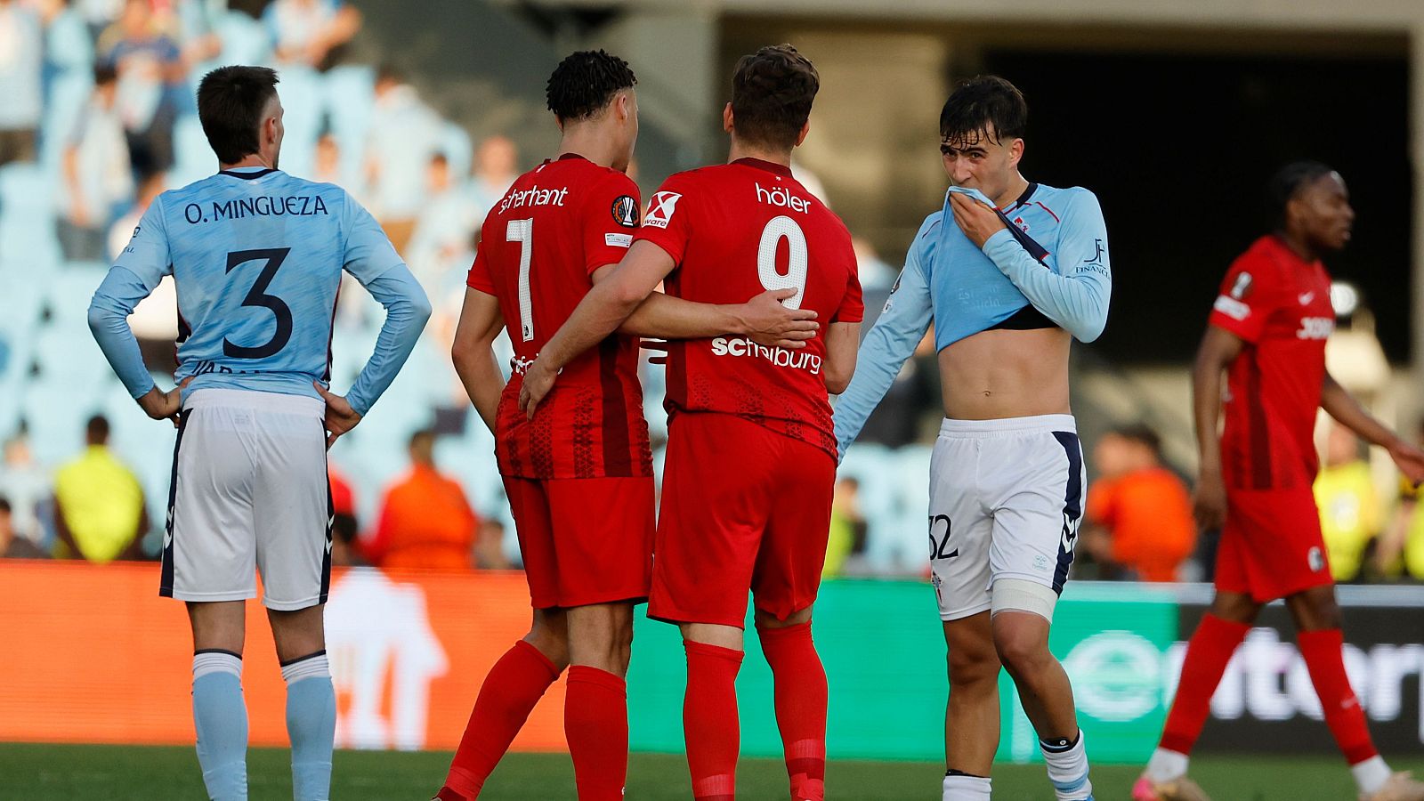 Jugadores de fútbol, vestidos con camisetas de color celeste y rojo, reaccionan en el campo. Un jugador del Celta de Vigo se cubre la cara con su camiseta, mientras que jugadores del Friburgo celebran un gol.