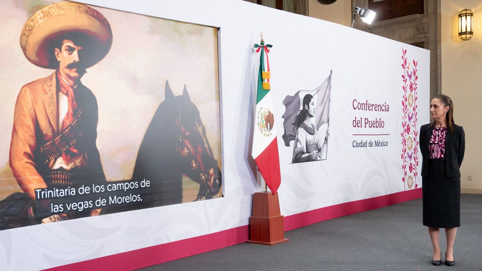 Claudia Sheinbaum, vestida con falda y chaqueta negra, ofrece una conferencia en Ciudad de México. Tras ella, un panel exhibe imágenes y textos, incluyendo la bandera de México y una imagen de Emiliano Zapata.