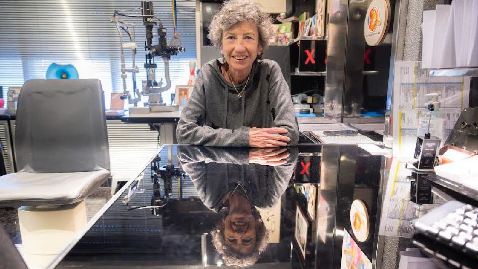 La doctora, amb cabells arrissats grisos, està asseguda darrere d'una taula reflectant, amb una sudadera grisa i una cadena. Darrere d'ella, es distingeix equipament mèdic i estanteries amb objectes.