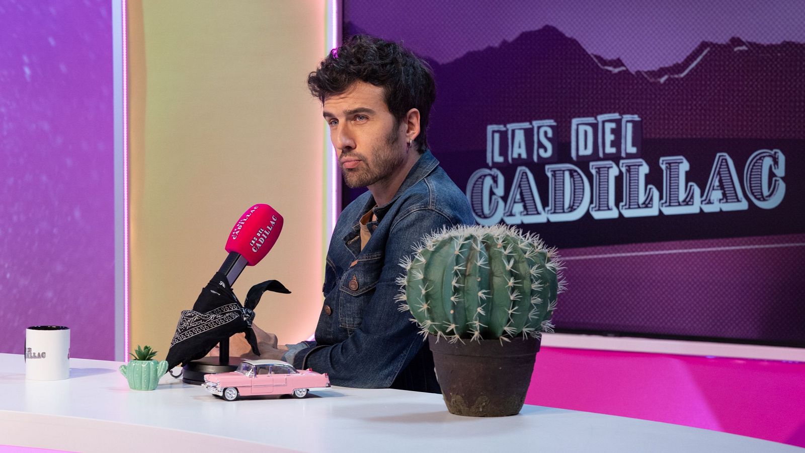 Guille Galván en "LAS DEL CADILLAC" con micrófono rojo, coche de juguete Cadillac rosa y fondo colorido.