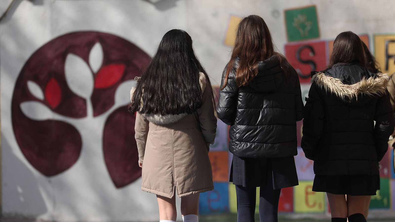 Fotografía de tres nñas de espaldas en el patio de un colegio vestidas con abrigo y el uniforme escolar
