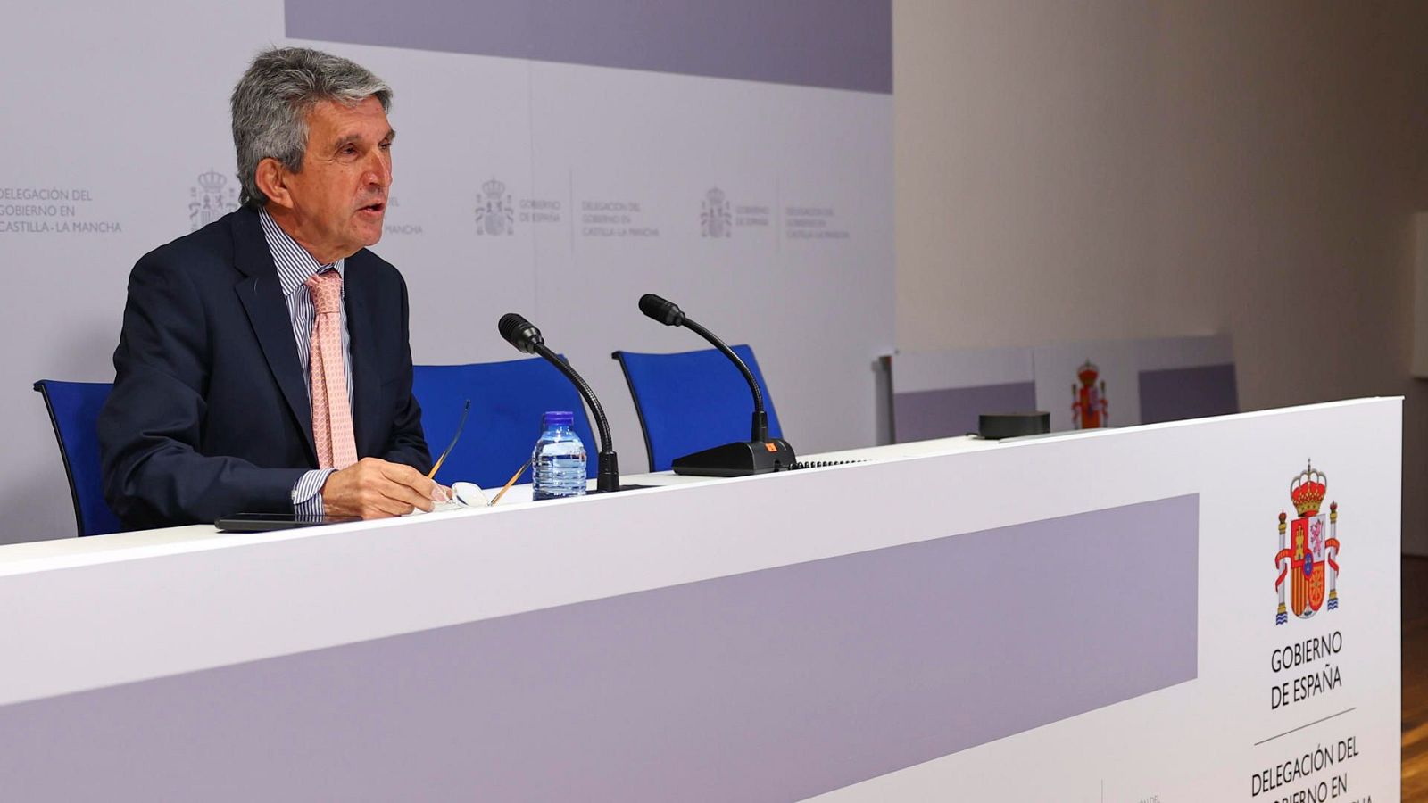 Un hombre con traje azul marino y corbata, posiblemente José Pablo Sabrido, se encuentra sentado frente a una mesa con micrófonos, una botella de agua y gafas. En el panel frontal de la mesa se observa un escudo y texto institucional.