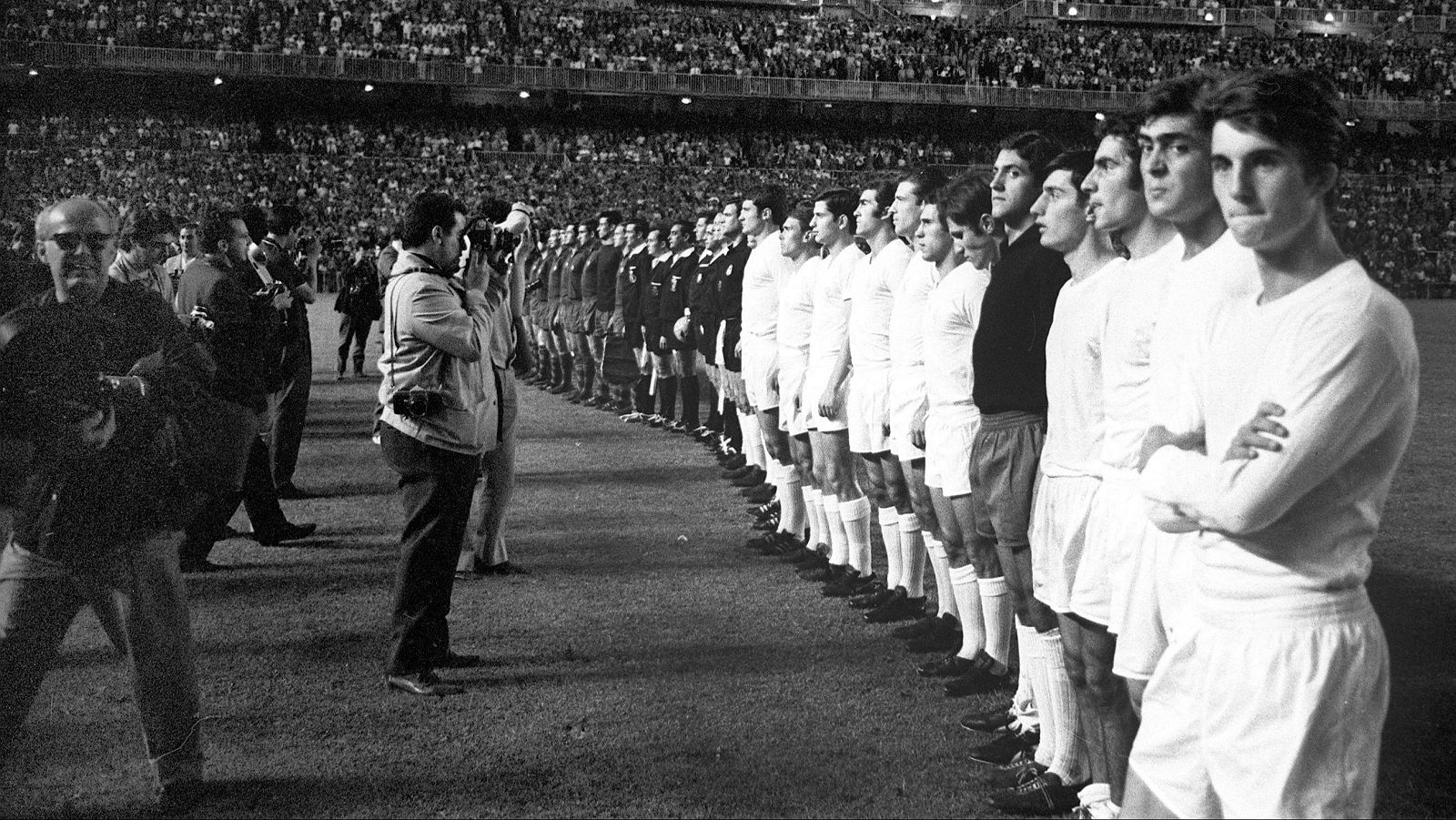 Dos equipos de fútbol, uno con uniformes claros y otro con oscuros, se enfrentan en un campo, con una multitud observando desde las gradas. Varios fotógrafos y personal de apoyo se encuentran en primer plano.