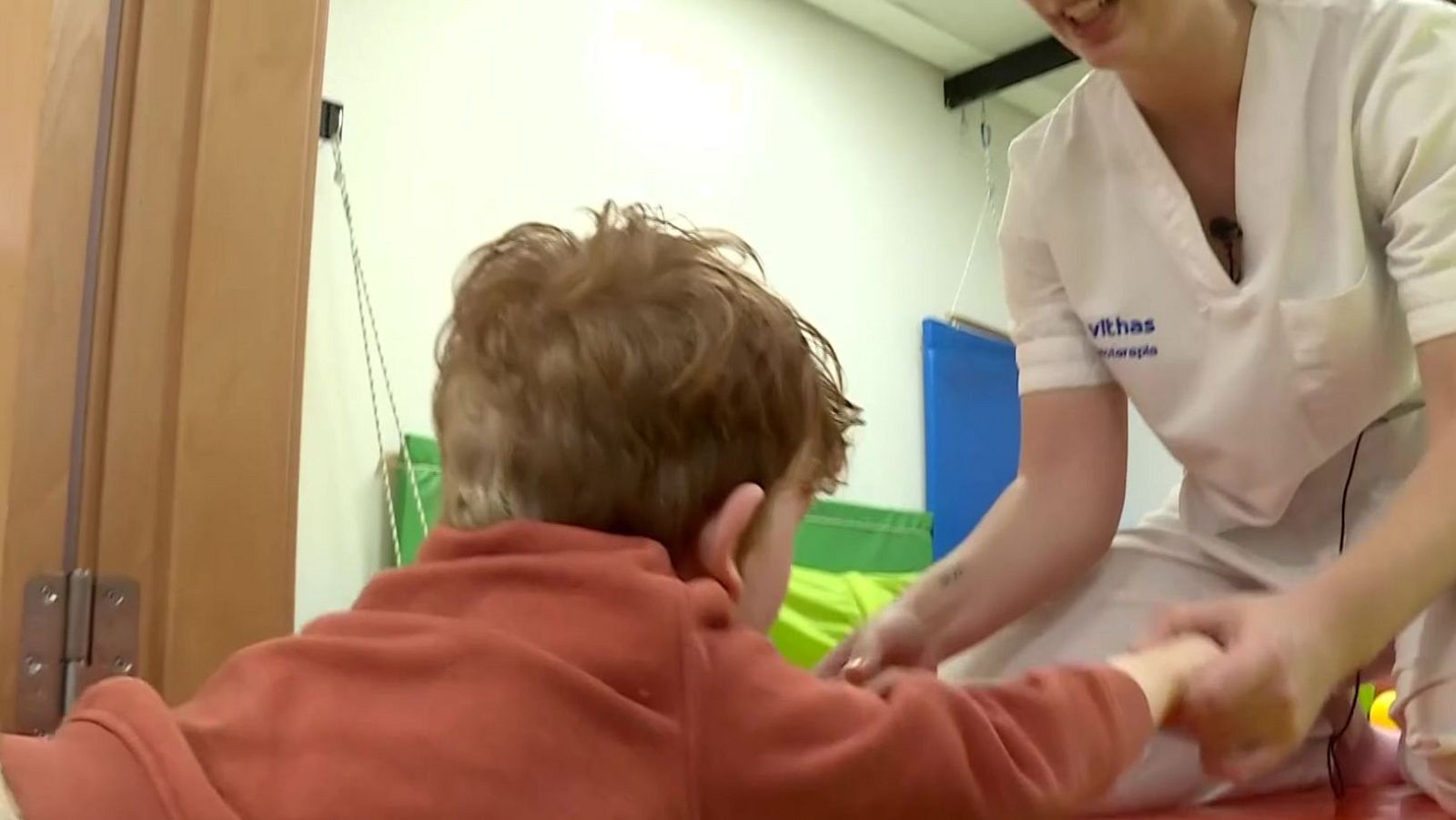 En una sala de terapia, un terapeuta con bata blanca interactúa con un niño pelirrojo, sosteniendo sus manos. Se observa equipamiento especializado en el fondo.