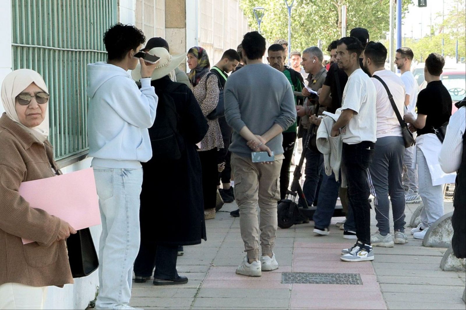 En la imagen, un grupo diverso de personas espera en la calle. Se distinguen individuos con vestimenta variada, como una mujer mayor con hijab y un joven con sudadera, así como personas de espaldas y otros jóvenes.