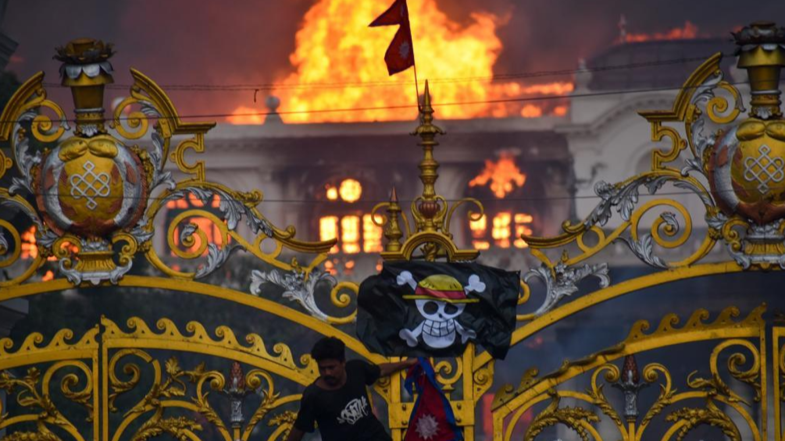 Un edificio en llamas es el escenario de un disturbio, con humo visible. Un hombre sostiene una bandera de Nepal y otra con una calavera, frente a una verja ornamentada.