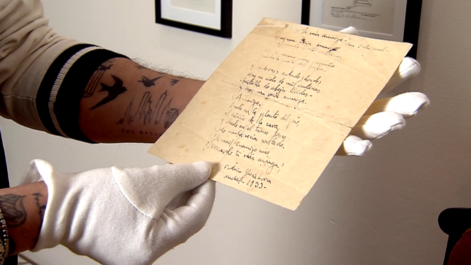 Unas manos con guantes blancos sostienen un manuscrito amarillento con texto escrito a mano. Se aprecian tatuajes en el brazo izquierdo, uno de ellos con golondrinas en vuelo.