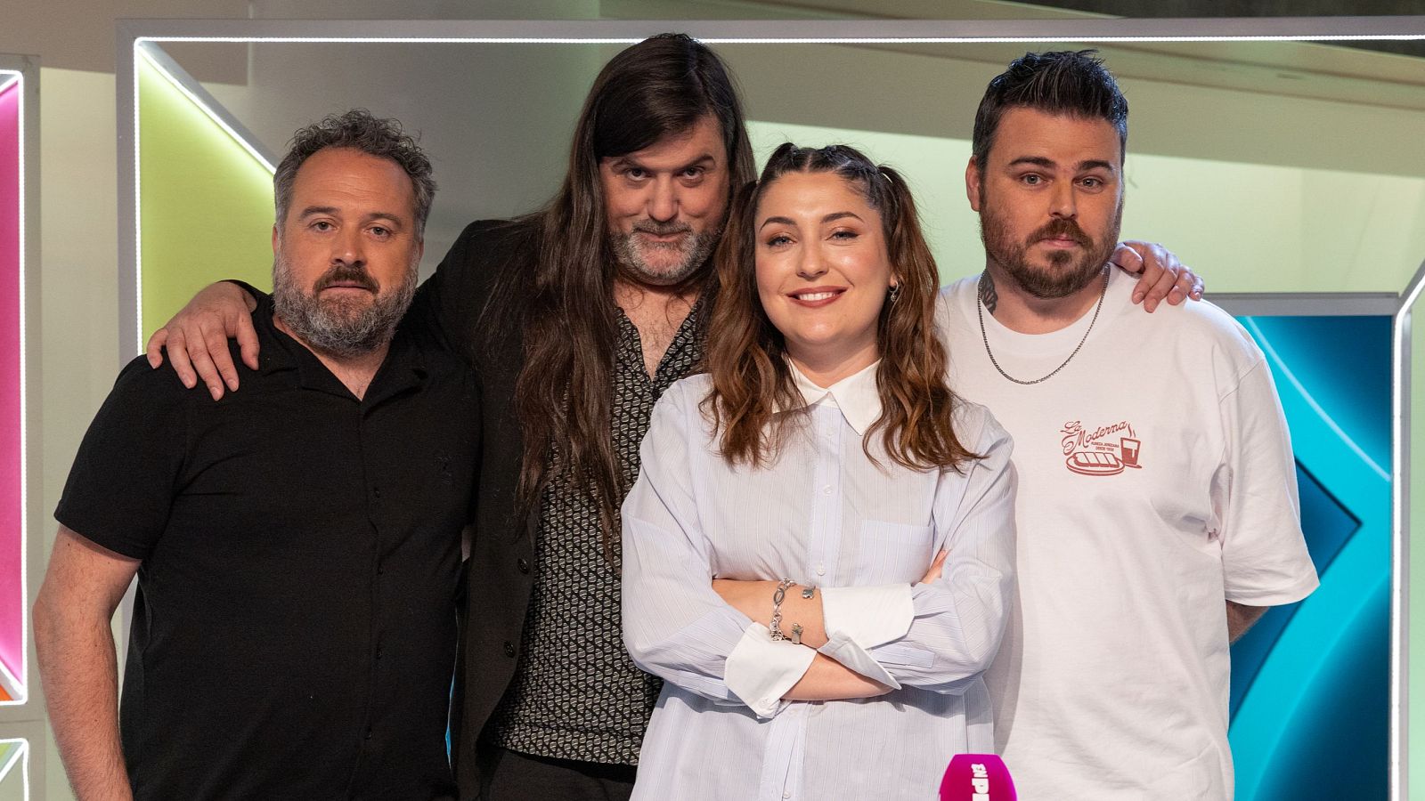Cuatro personas se encuentran sentadas en un banco al aire libre, con árboles y una torre de telecomunicaciones como telón de fondo. Los individuos visten ropa casual, como camisetas, chaquetas y sudaderas.