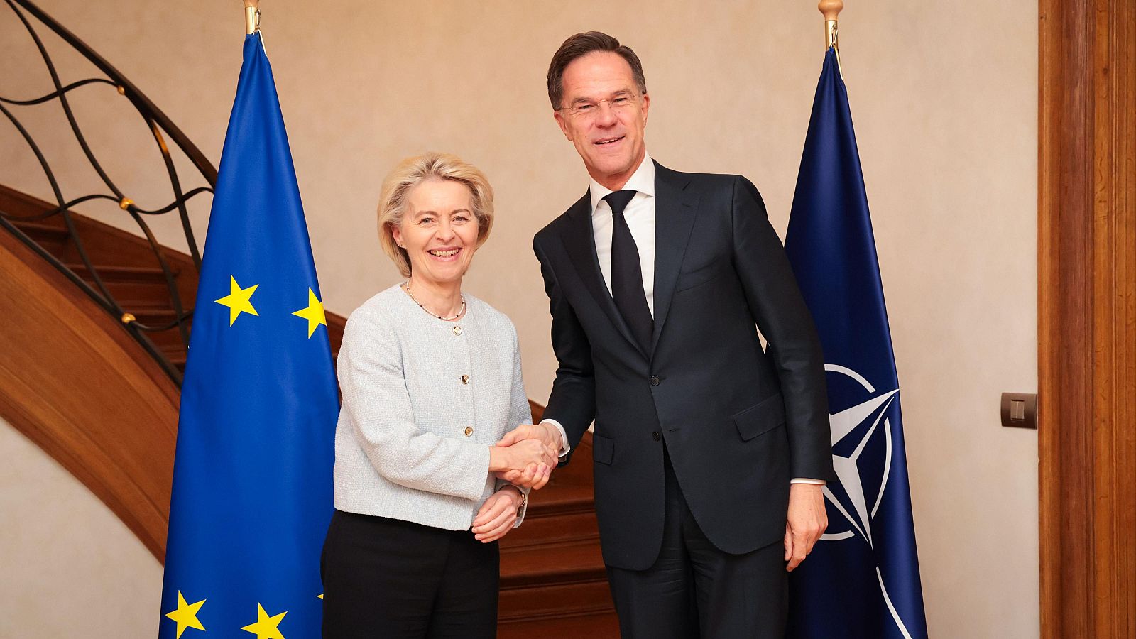 En Bruselas, Ursula von der Leyen y Mark Rutte se dan la mano, sonriendo. La presidenta de la Comisión Europea viste una chaqueta clara, mientras que el secretario general de la OTAN lleva un traje oscuro. Al fondo, se aprecian banderas.