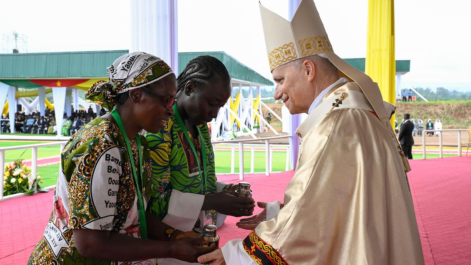 El Papa, con vestimenta ceremonial, interactúa con dos mujeres africanas durante una misa en un evento al aire libre, posiblemente en Camerún. Las mujeres visten ropas tradicionales con estampados vibrantes, mientras que el fondo muestra una multitud y decoraciones.