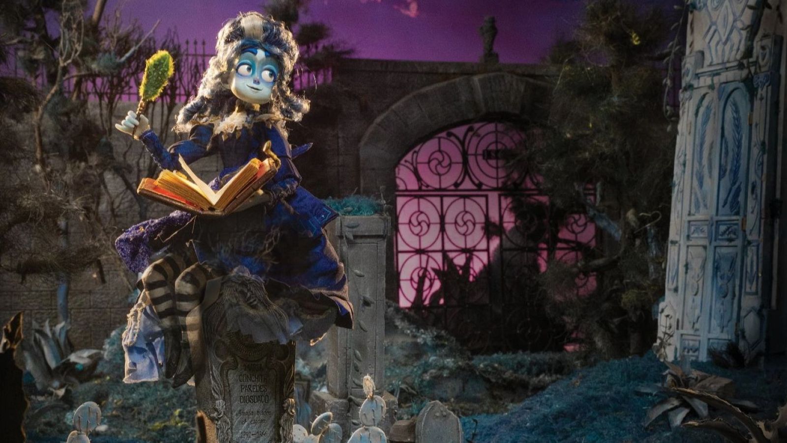 Una muñeca de aspecto fantasmal, con piel pálida y cabello rizado, está sentada sobre una lápida en un cementerio. Viste un vestido oscuro y sostiene un espejo y un libro abierto, en un ambiente sombrío con lápidas y una puerta de hierro.