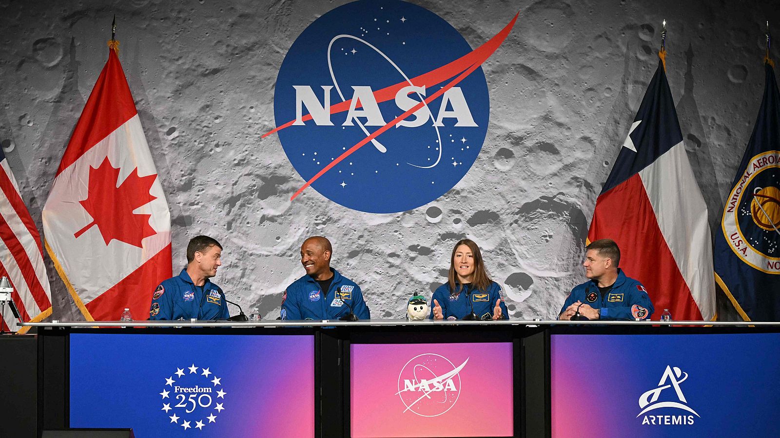 Cuatro astronautas, vestidos con chaquetas azules con parches, se encuentran sentados detrás de una mesa durante una conferencia. El fondo muestra un diseño que simula la superficie lunar, con logos y banderas.