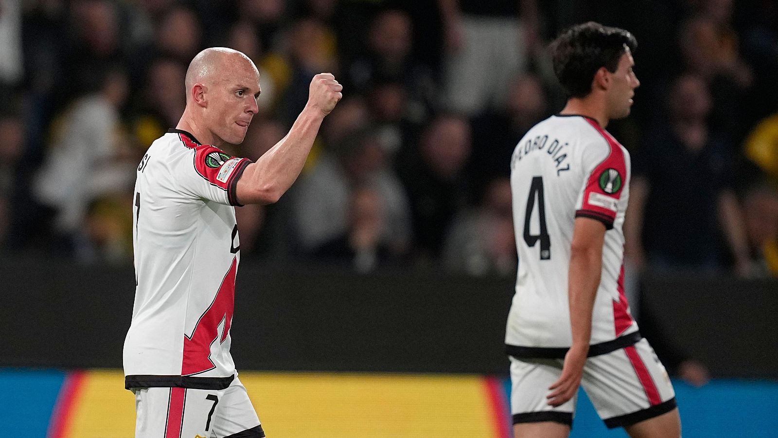 Isi Palazón, con la camiseta número 7, celebra un gol junto a otro jugador con la camiseta número 4, ambos vistiendo la equipación blanca y roja de su equipo en un estadio lleno de público.