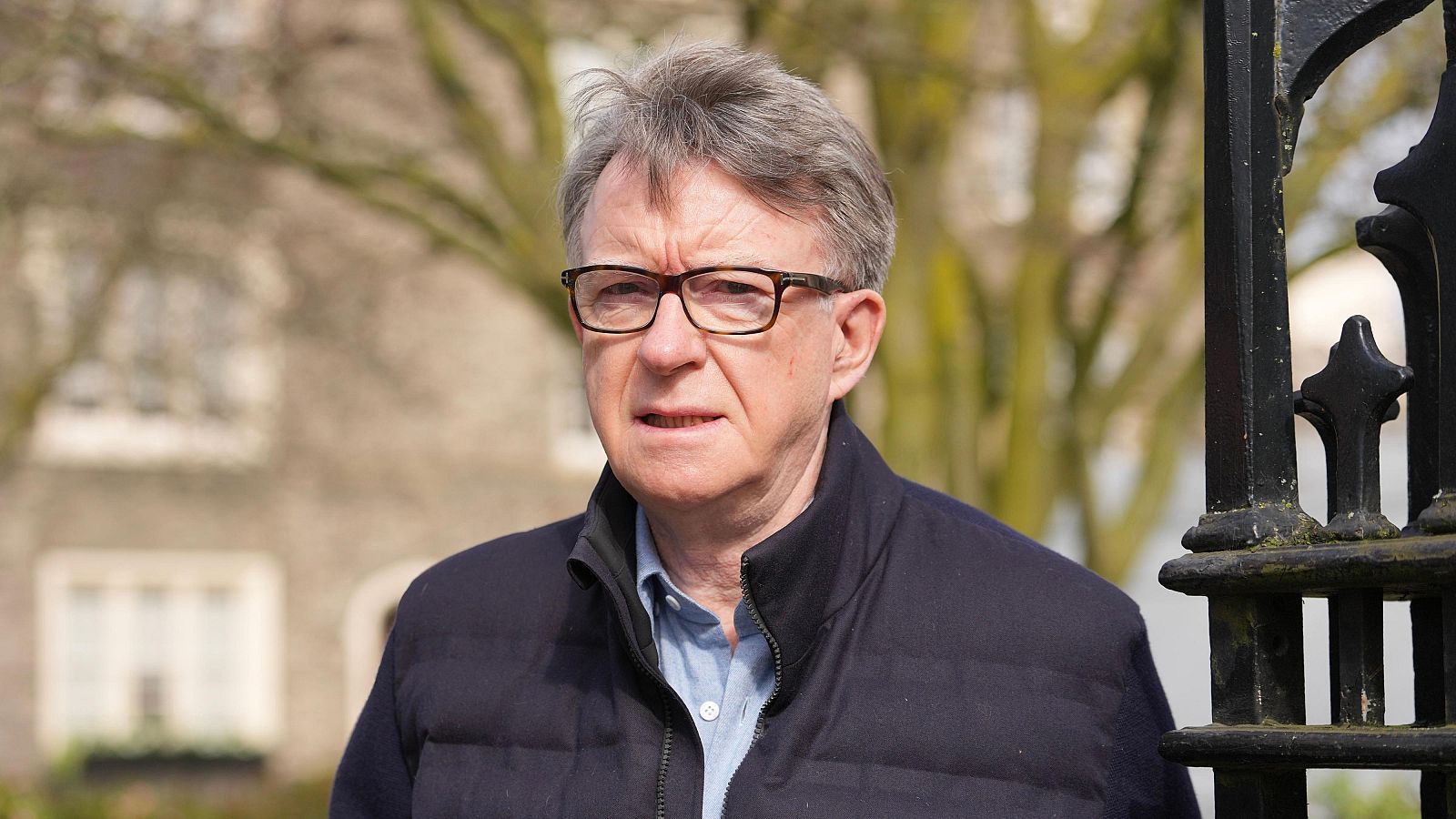 Peter Mandelson, con camisa clara y chaqueta oscura, se encuentra frente a una reja de hierro, con un edificio de piedra y árboles sin hojas al fondo, posiblemente en una estación fría.