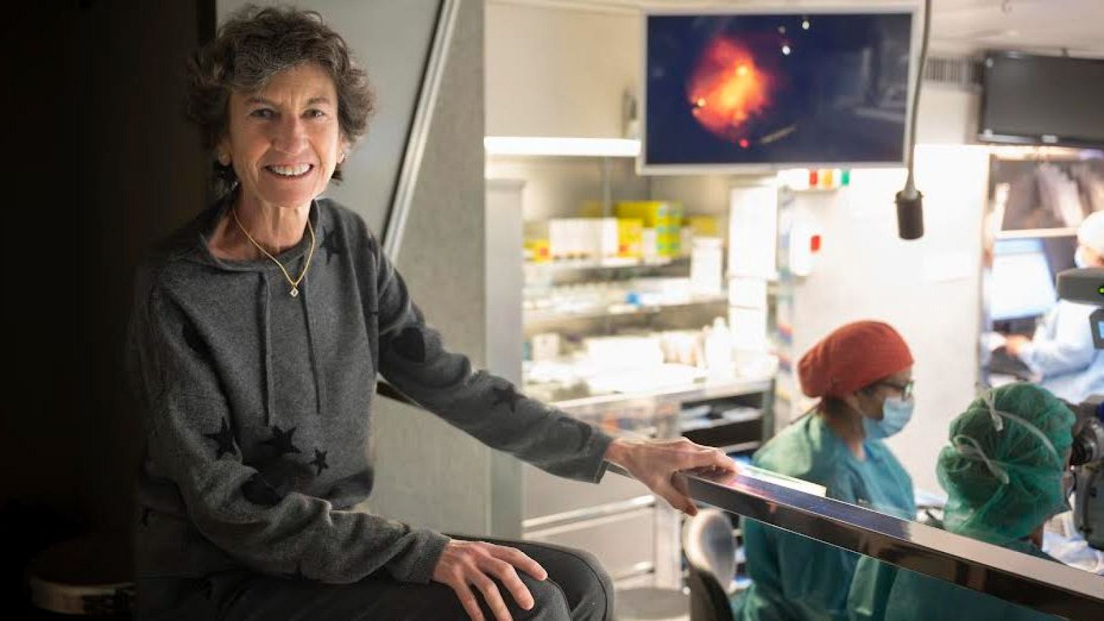 En una sala mèdica, la doctora Elena Barraquer somriu mentre està asseguda. Porta una dessuadora grisa amb estrelles i un collaret daurat, amb personal sanitari al fons.