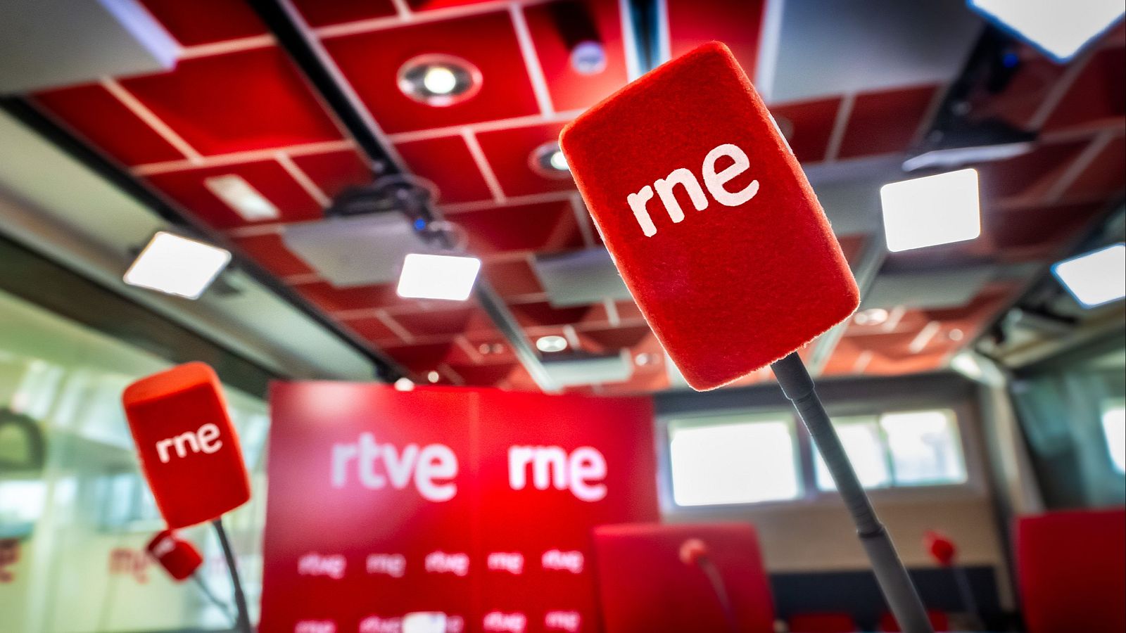 Un estudio de radio con micrófonos rojos y el logotipo de "rne". Se observa un panel rojo con los logotipos de "rtve" y "rne", y el techo presenta patrones geométricos.