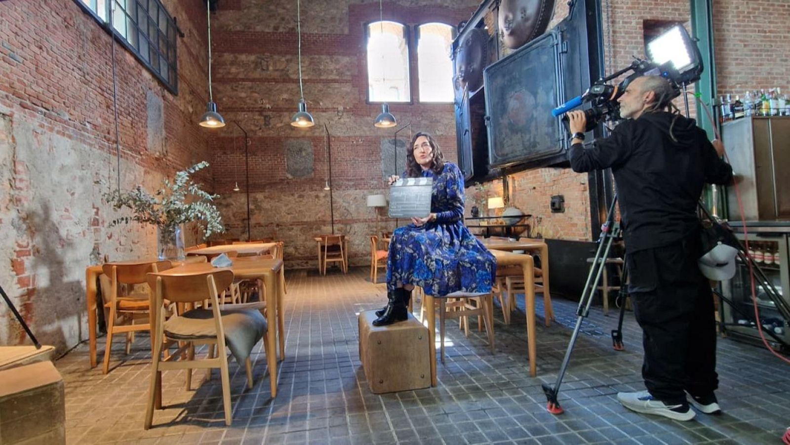 En un espacio con paredes de ladrillo visto, una mujer con un vestido azul y botas negras sostiene una claqueta mientras un hombre la graba con una cámara profesional. El entorno parece ser un restaurante o un lugar similar con mesas y sillas de madera.
