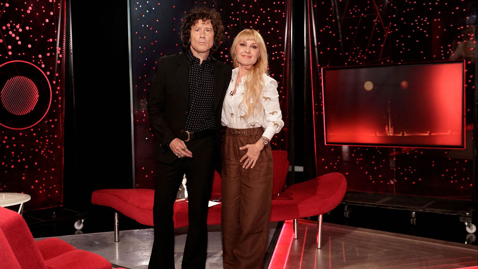 Bunbury, vestido con un traje negro y camisa de lunares, posa junto a una mujer rubia con una blusa estampada, en un estudio de televisión con sofás rojos y una pantalla al fondo.