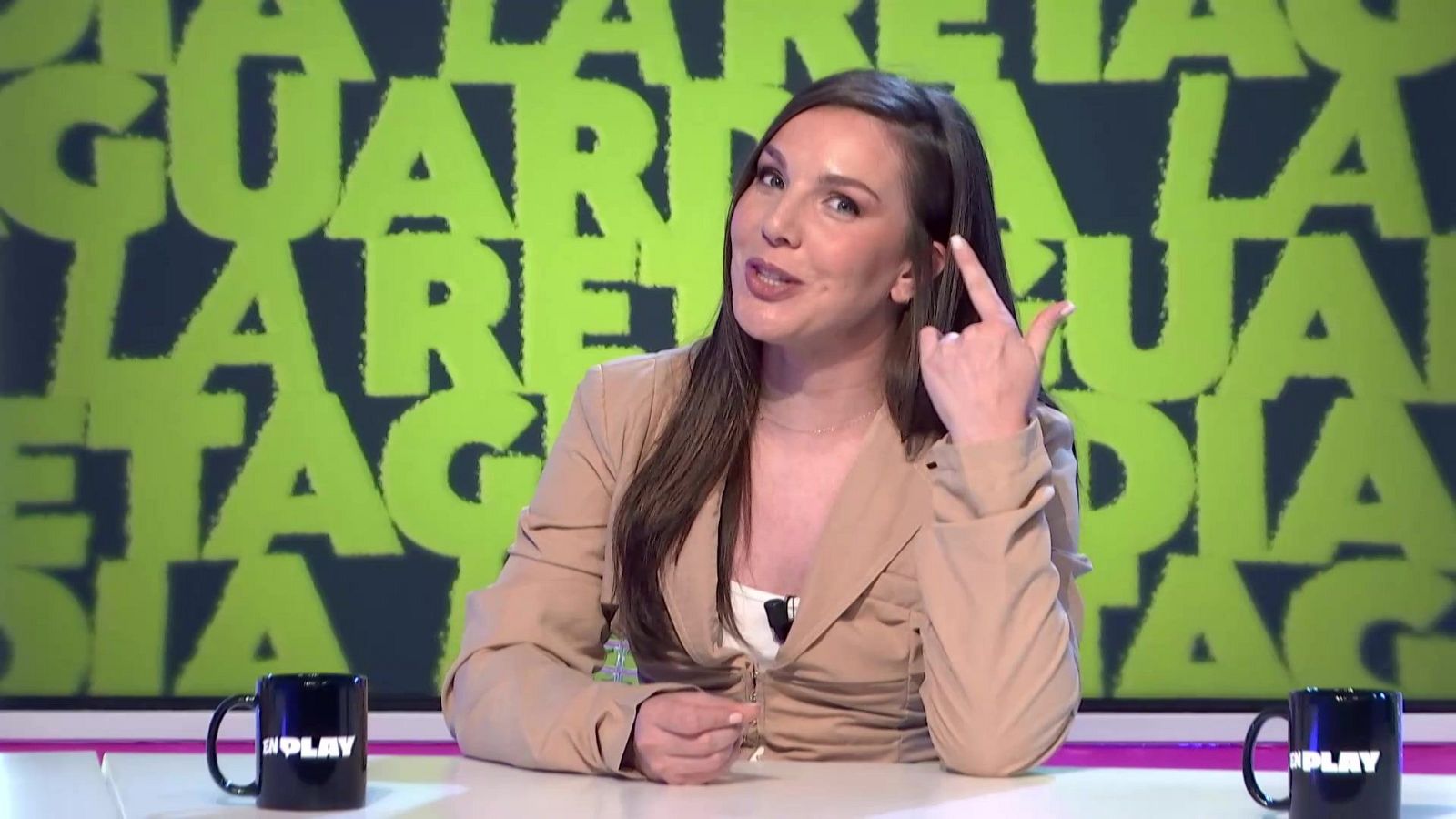 En la imagen, una mujer con chaqueta beige y cabello castaño señala su cabeza mientras está sentada frente a una mesa. El fondo muestra la repetición de un texto en letras verdes, y sobre la mesa hay dos tazas negras con un logo blanco.