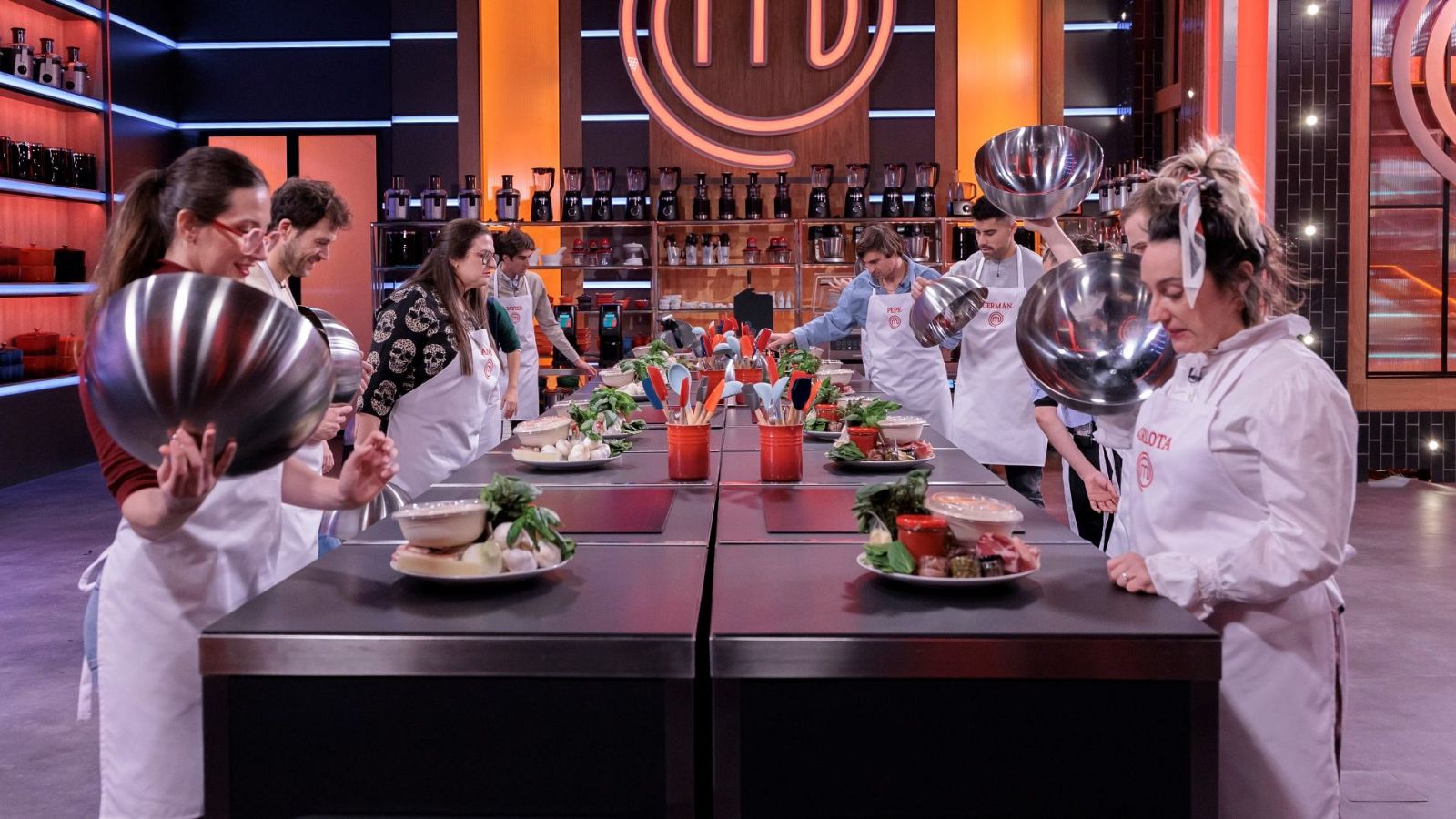 En una cocina profesional, un grupo de aspirantes a chef, con delantales blancos, se preparan para cocinar. Sobre la mesa, se observan ingredientes frescos y utensilios, posiblemente para la elaboración de pizzas.