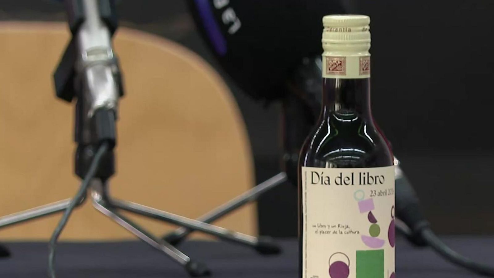 Una botella de vino tinto con una etiqueta que celebra el "Día del Libro" del 23 de abril de 2023, con un diseño abstracto y la frase "Libros y un Rioja, el placer de la cultura", situada frente a dos micrófonos.
