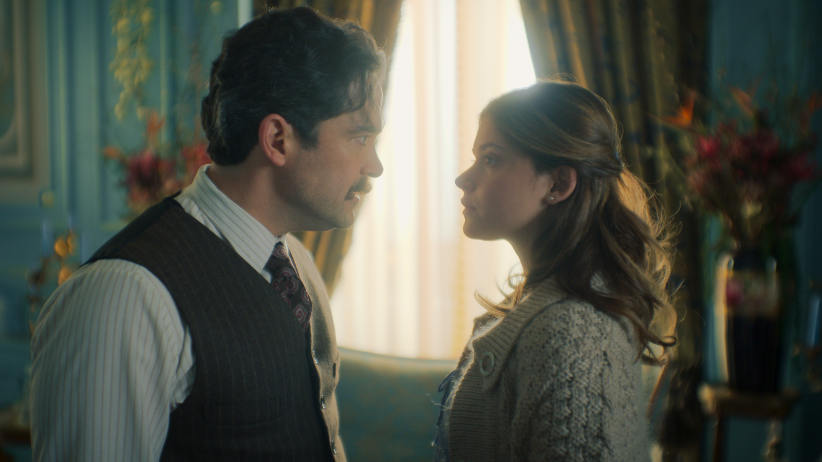 Un hombre con camisa a rayas y chaleco, junto a una mujer con cárdigan, se miran en una habitación con cortinas claras y muebles clásicos, iluminada por la luz de una ventana.