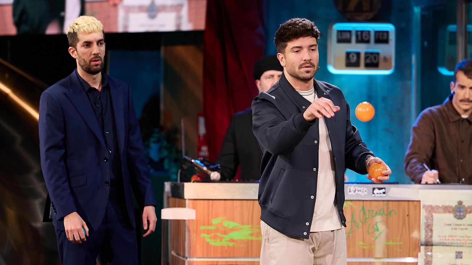 David Broncano y Pablo Alborán compiten en un juego de petanca en un plató de televisión. Broncano, con el pelo teñido, observa a Alborán lanzar una bola naranja, mientras un hombre escribe y un marcador de fecha es visible.