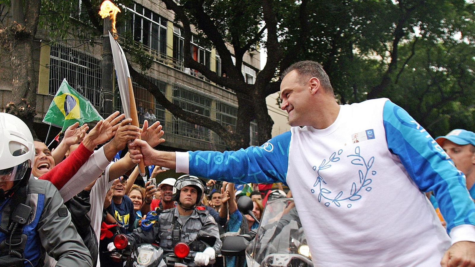 Oscar Schmidt, leyenda del baloncesto, sonríe mientras porta la antorcha olímpica, extendiéndola hacia una multitud que busca tocarla, con banderas y motocicletas de policía presentes en la escena.